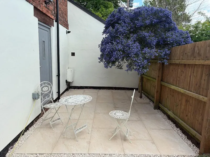 Compact square of large light beige tiles with white gravel borders, a metal bistro set and purple flowering shrub over a wooden fence.