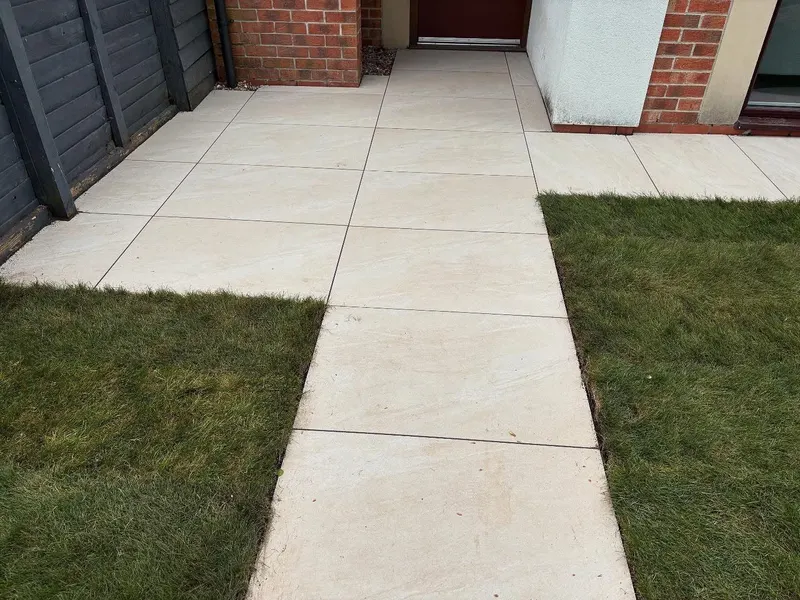 Light square paving in a central walkway and side pad between strips of new grass, leading to a dark door on brick and render walls.