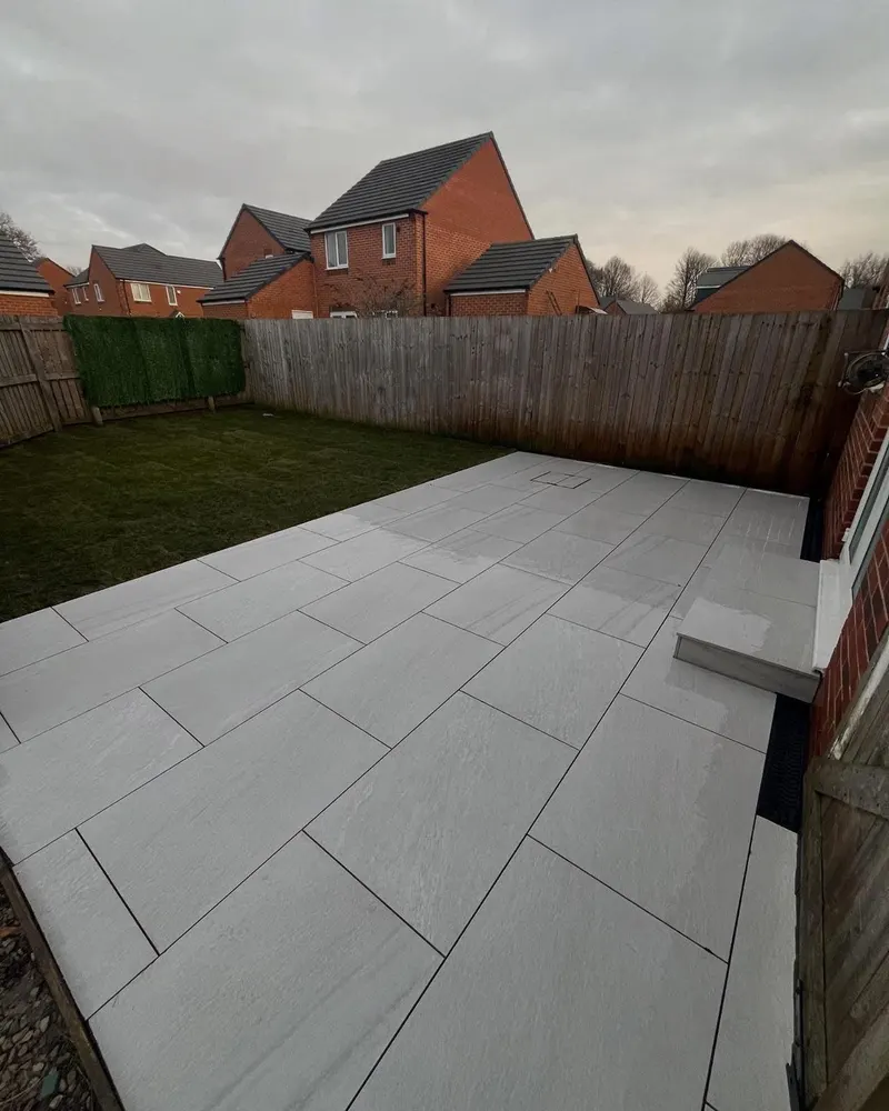 Large light grey staggered slabs with dark joints, integrated manhole cover and a small step to the house, beside lawn and panel fencing.