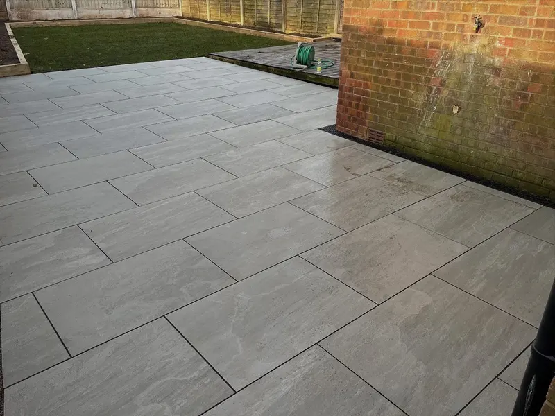 Light grey flagstones in running bond from a brick wall with channel drain, past sleeper-edged lawn to a dark stained decking area.