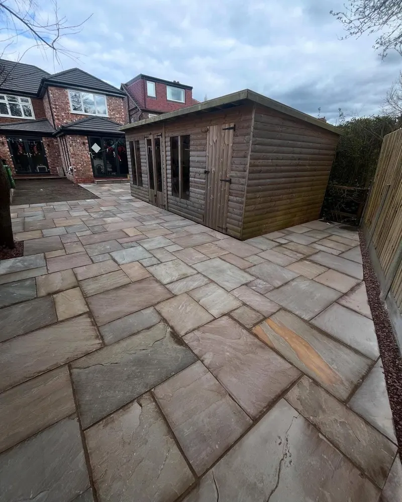 Multi-coloured riven stone patio wrapping a log-cabin style wooden garden room, bordered by dark gravel against a tall fence.