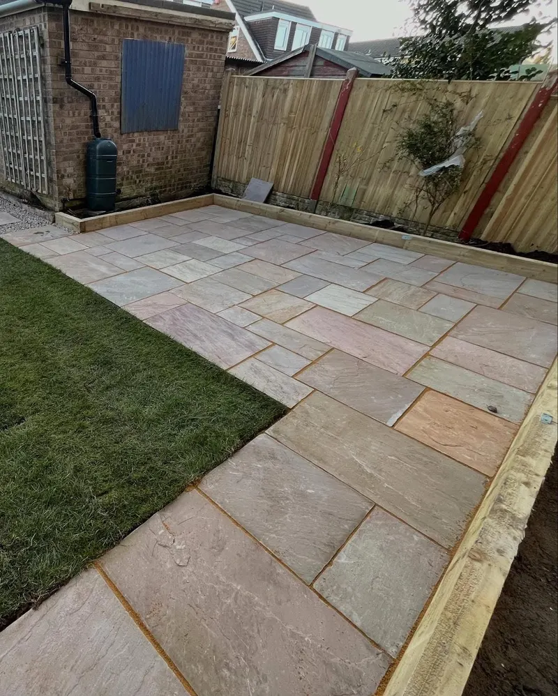 New riven stone flags in buff, grey and pink tones beside fresh turf, framed by timber sleepers and a wooden fence with a water butt.