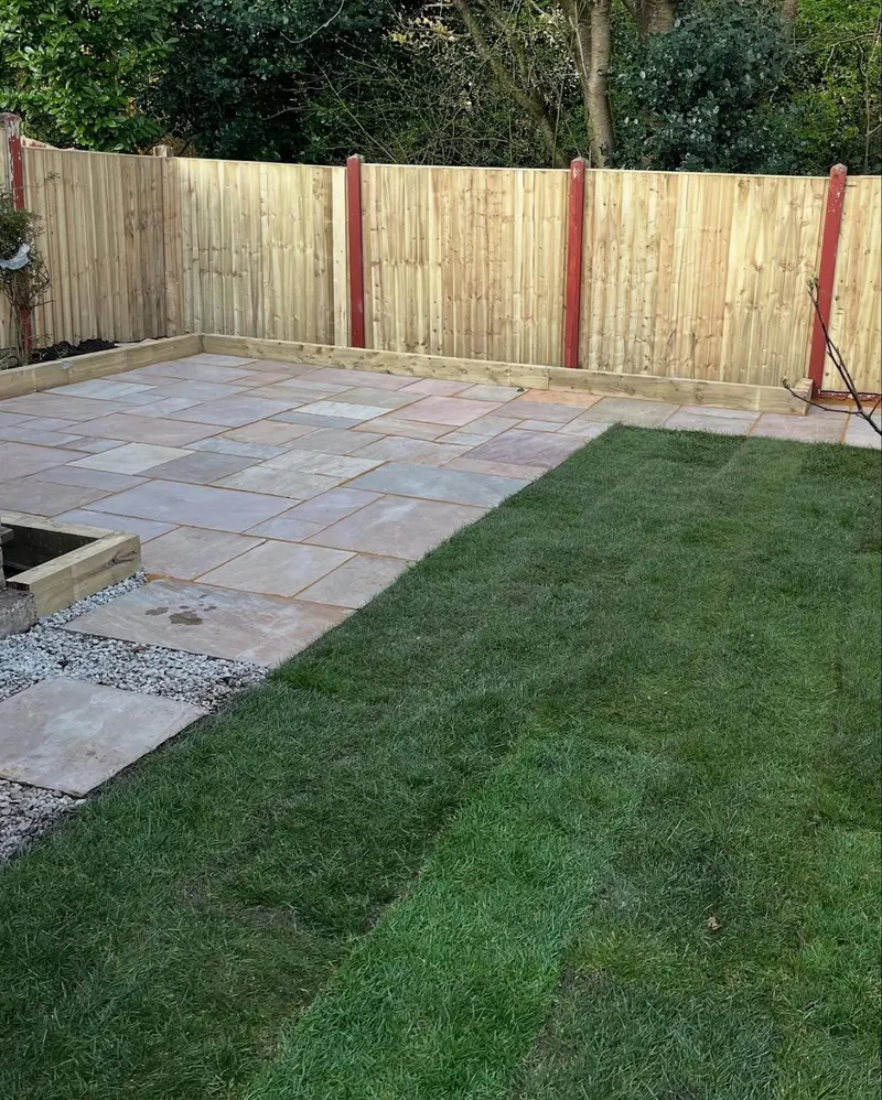 Rectangular patio of mixed-size sandstone slabs in warm tones, sleeper borders, new green turf and a light wooden fence with brown posts.