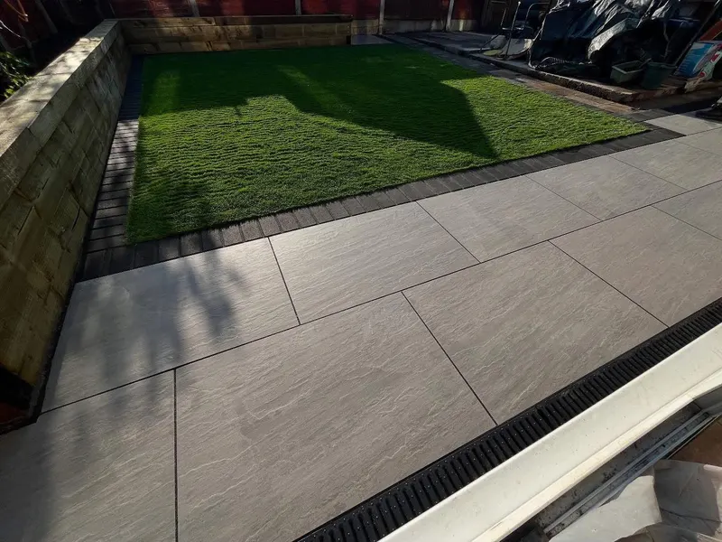 Light grey textured slabs next to a lawn edged with timber sleepers, separated by a row of small dark pavers and a channel drain at the threshold.