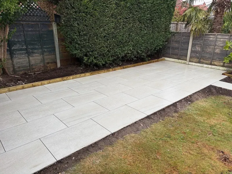 Large rectangular pale paving in staggered bond with a timber sleeper edge, hedge and fencing around a recently laid lawn border.