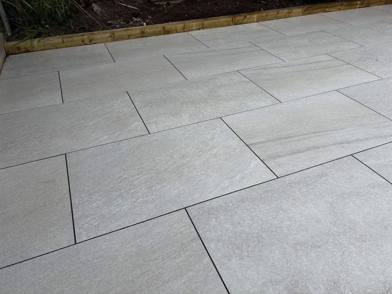 Detail of light grey textured rectangular pavers in running bond with charcoal grout lines, meeting a timber sleeper and planting bed.