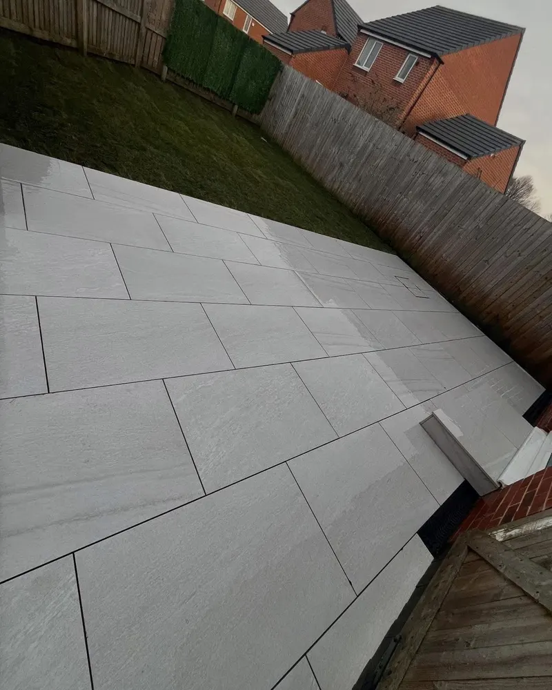 Expansive light grey staggered paving beside lawn and fence, including a recessed utility cover paved to match the surrounding slabs.