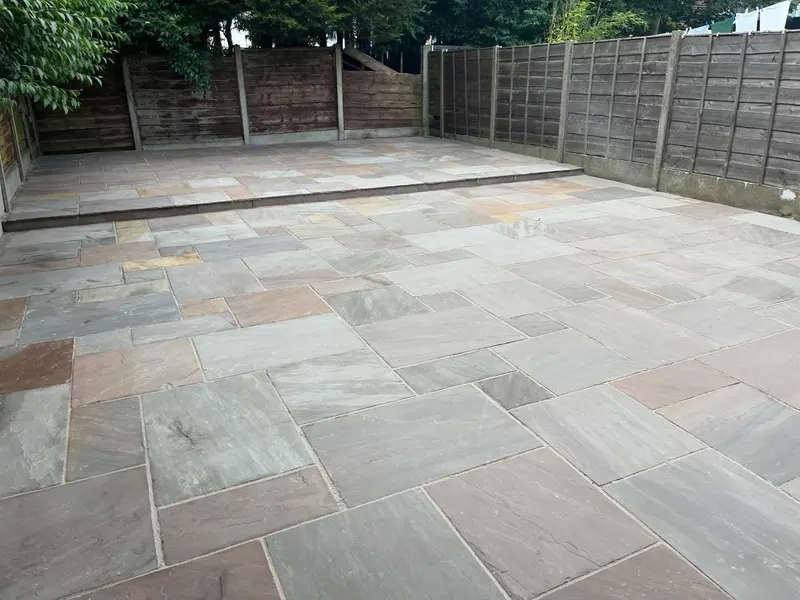 Lower and upper paved levels of multi-tonal rectangular stone flags linked by a single stone step, enclosed by a wooden lap fence.