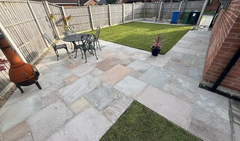 Large mixed-tone flagstone patio wrapping a central lawn, with metal table and chairs, chiminea, drainage channel along the brick wall and wooden fencing.