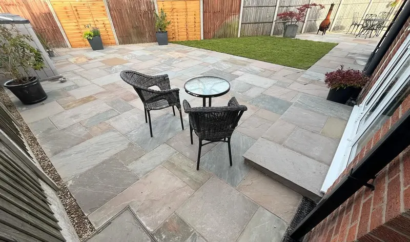 Varied grey and buff flags with a single step to glass doors, wicker bistro set, potted plants and gravel strip beside a panel fence.