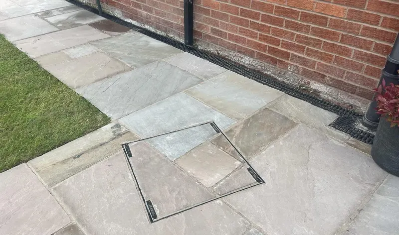 Natural stone flags in grey and buff beside lawn and brick wall, with a square manhole infilled in matching stone and a channel drain.