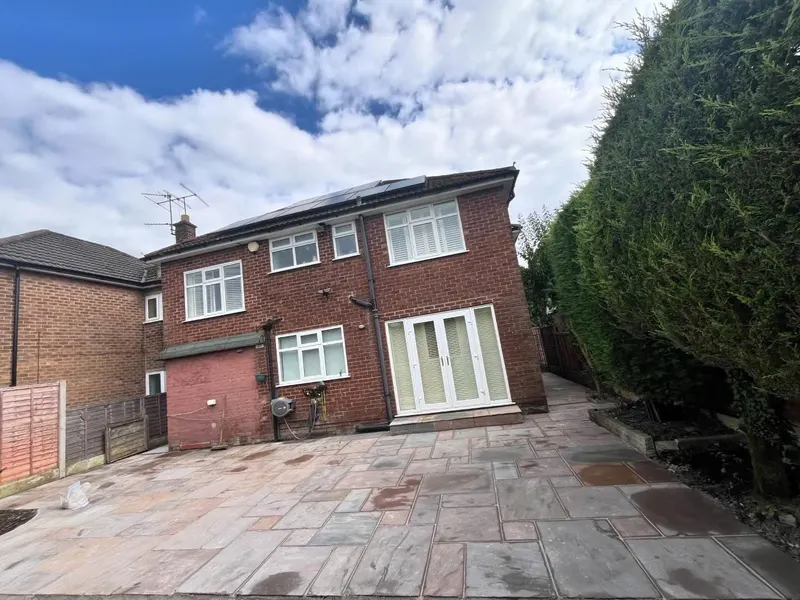 Full-width backyard paved in large mixed-colour stone slabs from white French doors outward, bounded by fence, hedge and a small brick outbuilding.