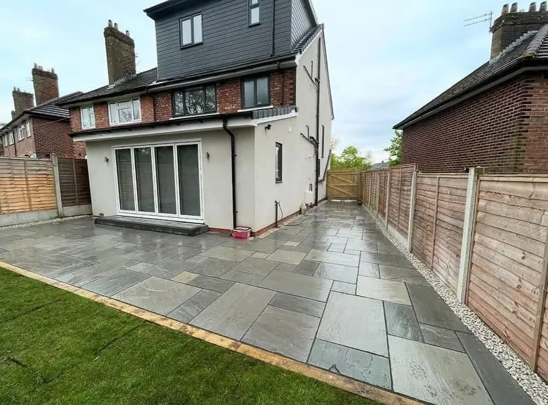 Modular grey riven slabs across the rear and side passage of a rendered extension, with bi-fold doors, lawn sleeper edge and gravel by the fence.