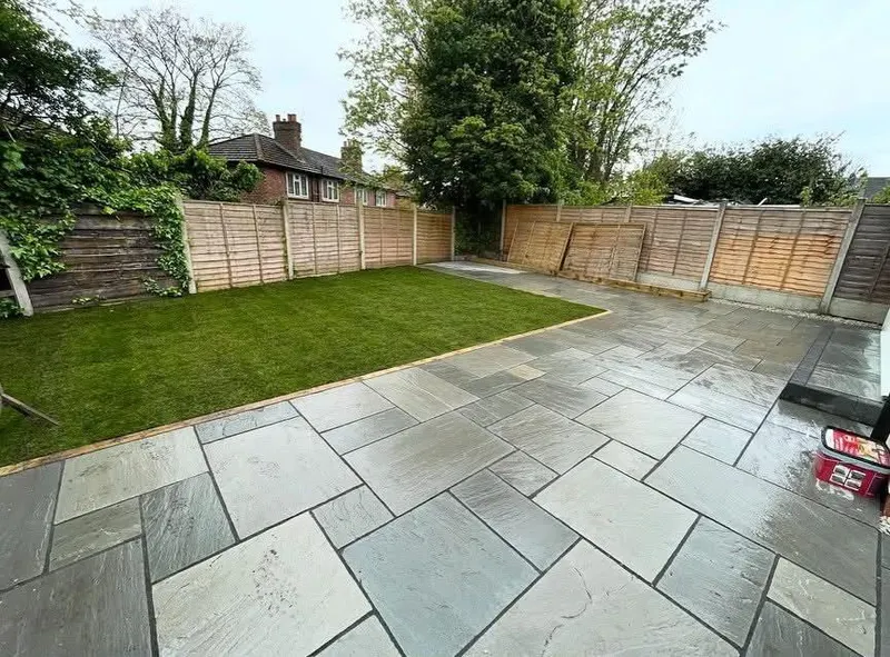 Wet-looking grey stone flags in varied sizes beside a rectangular grass area, timber edging, wooden fence and ivy on the boundary.