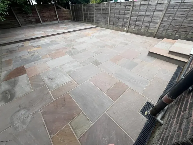 Wide backyard paved in mixed grey and buff slabs with two stone steps, a raised tier, slot drain along the brick wall and lap panel fencing.