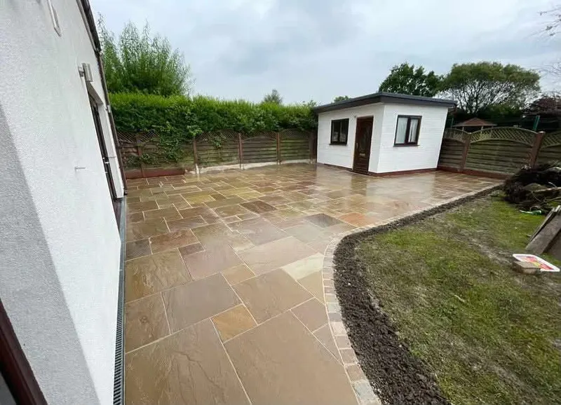 Large buff and grey stone patio sweeping to a lawn with a small cobble sett border, white garden room ahead and hedge behind decorative fencing.