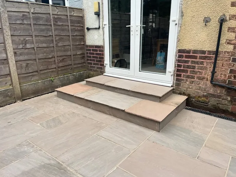Matching riven stone flags forming a double step up to white French doors on rendered and brick walls, with fence and channel drain visible.