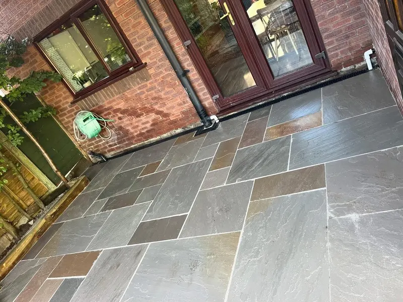 Dark and light grey riven slabs with light pointing against a brick wall, dark French doors, channel drain and hose reel mounted above.