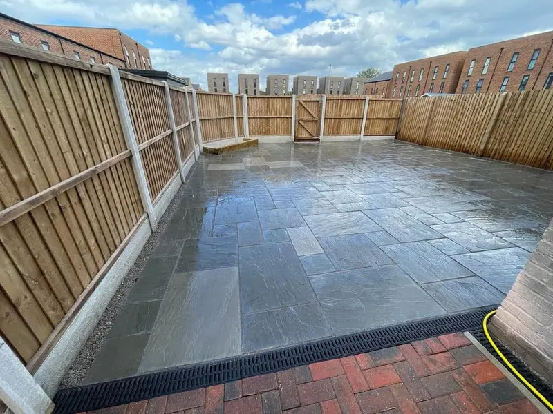 Spacious grey riven patio meeting a short stretch of red and charcoal block paving, black linear drain, wooden gate and gravel fence strip.