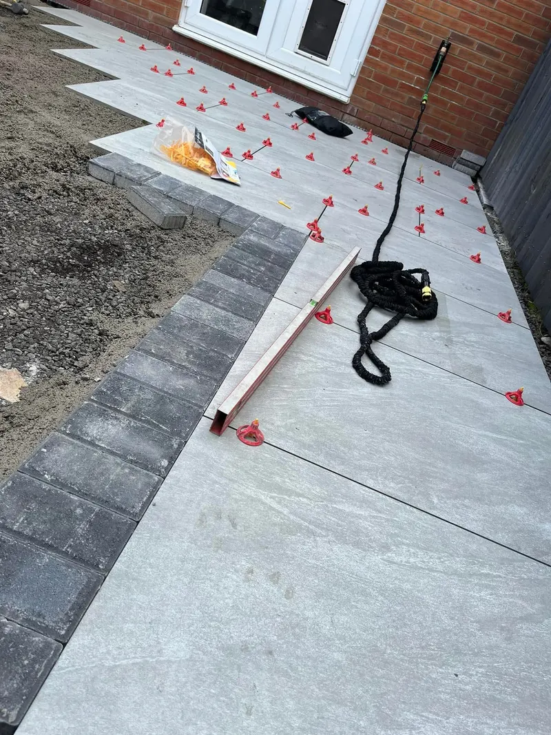 Work in progress showing large light grey slabs with levelling clips beside a charcoal brick border row, spirit level and hose on the finished section.
