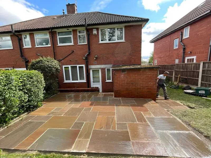 Freshly laid multi-size sandstone flags in tan and grey behind a red brick house, with grass edge, hedge, fence and installer on site.