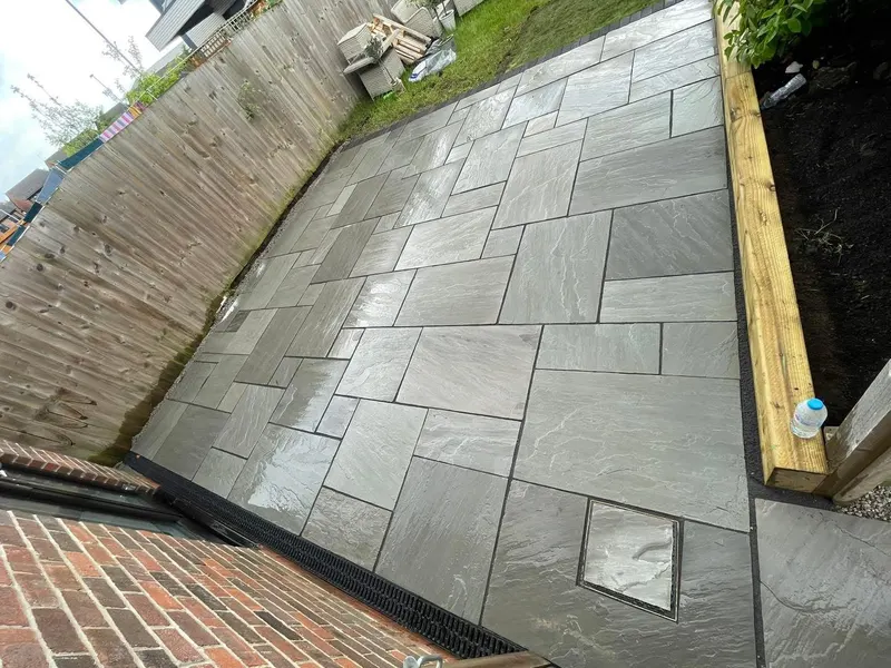 Overhead view of grey rectangular flags with inset manhole cover, channel drain on brick wall and timber sleeper forming a raised bed border.