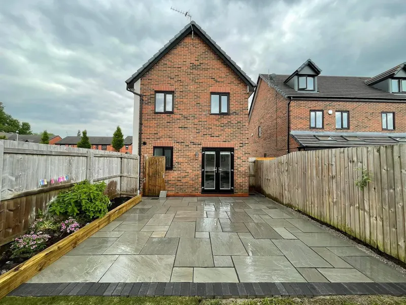 Large mixed grey and buff riven slabs across a fenced backyard, edged with dark rectangular blocks and sleeper planting bed by the brick house.