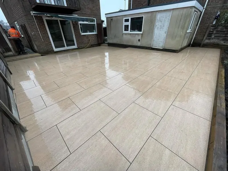 Wide light beige staggered paving from sliding doors along a red brick house to a rendered outbuilding, bounded by fence and sleeper wall.