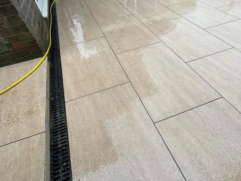 Close-up of large cream pavers in running bond beside a brick wall, with a continuous black slot drain and wet reflections on the surface.