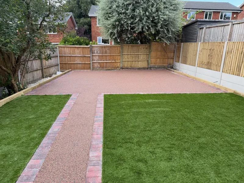 Rose-buff resin bound patio and connecting path bordered by mixed red and charcoal block pavers, flanked by bright green lawns and wooden fencing.