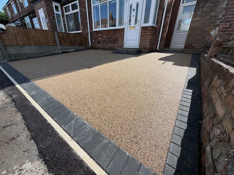 Buff-coloured resin bound driveway edged with dark block paving in front of a red brick house with a white bay window and porch.