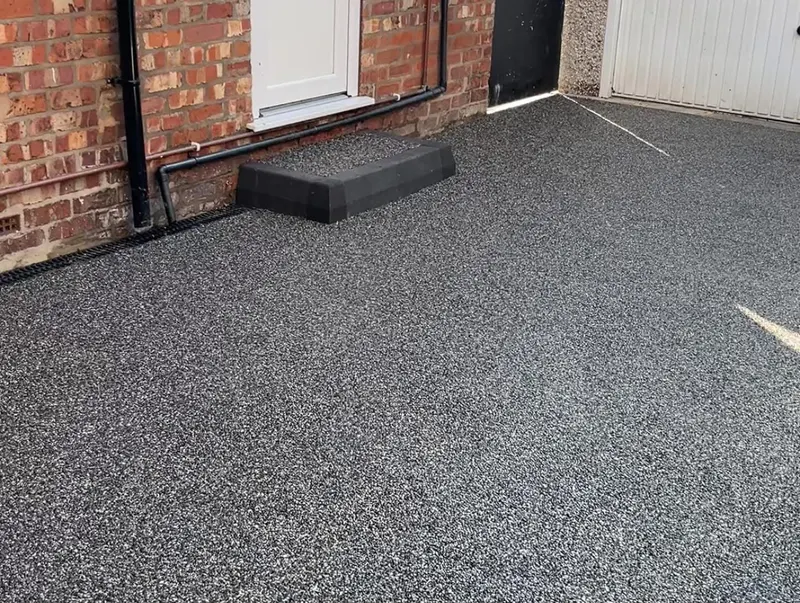 Salt-and-pepper grey resin bound courtyard with a small block-edged step to a white door, channel drainage, and a white garage door nearby.