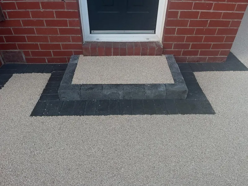 Raised entrance step surfaced in light beige resin and trimmed with charcoal block pavers, set against a red brick wall and dark front door.