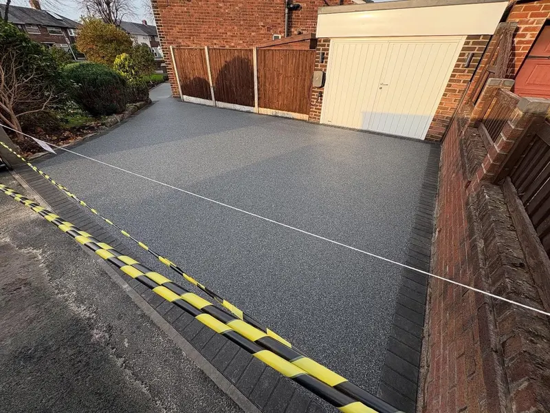 Silver-grey resin bound driveway with a charcoal block border leading to a white ribbed garage door, cordoned with hazard tape.