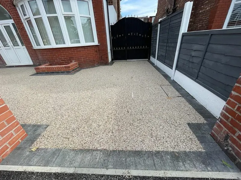 Light cream resin bound front driveway bordered by charcoal blocks, with a small brick planter and a block-framed manhole cover beside tall black gates.
