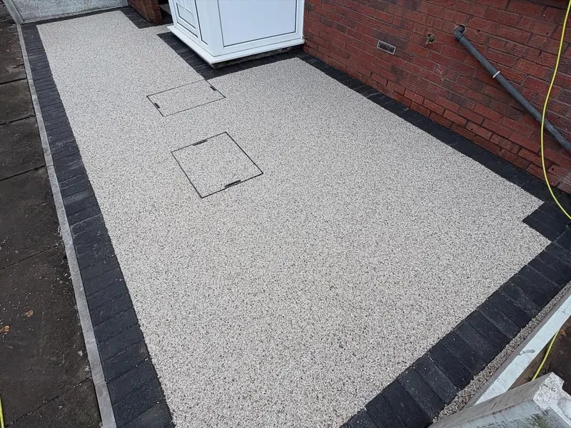 Light grey resin bound side area with two recessed covers filled to match, edged in dark block paving beside a red brick wall.