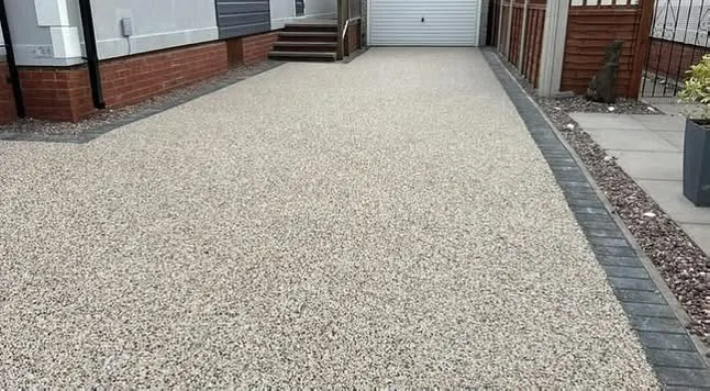Long narrow buff resin bound driveway with charcoal block edging running between a white-clad house and a brown fence toward a white garage.