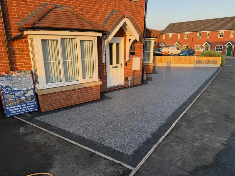 Light grey resin bound driveway with dark block borders shaped around a red brick house bay window and porch.