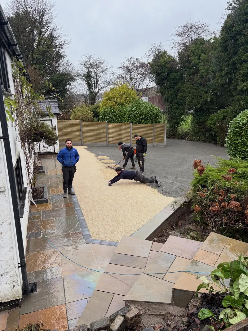 Workers smoothing a light tan resin bound layer next to a charcoal block border and existing multi-colour stone patio paving.