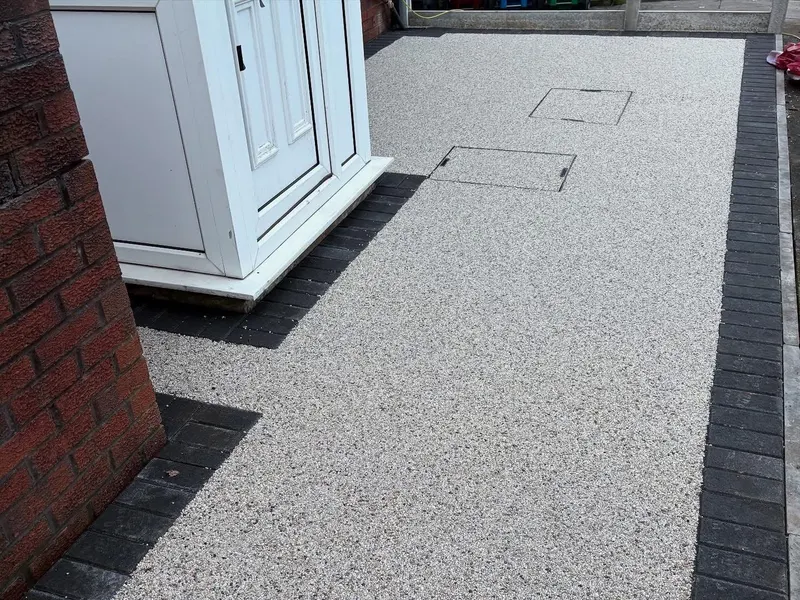 Close view of speckled light grey resin bound paving with dark block borders framing two flush manhole covers beside a white door and brick wall.