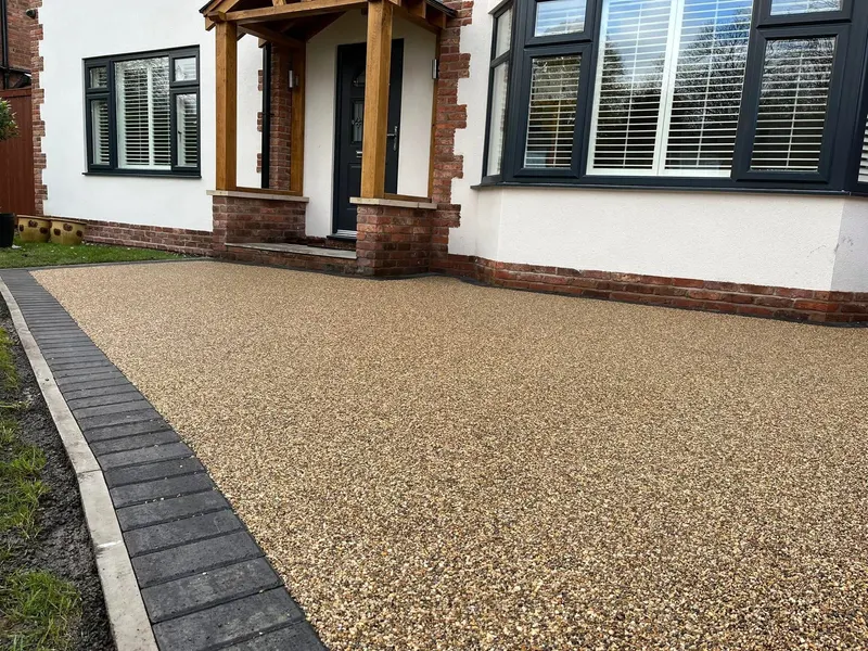 Golden-buff resin bound driveway with a charcoal block border along one edge, leading to a white-rendered house with a timber porch and black-framed windows.