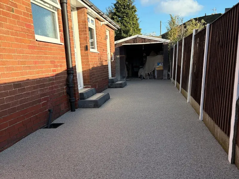 Narrow light grey resin bound path running between a red brick house with stone steps and a wooden fence toward a small garage.