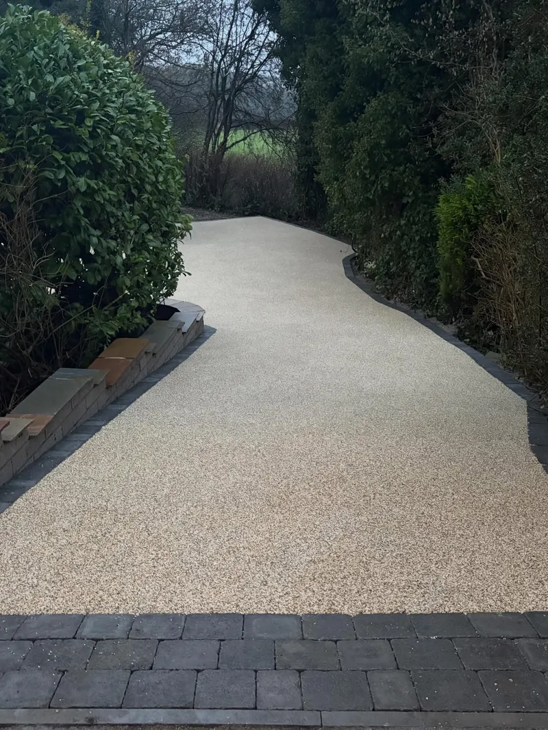 Gently curving cream resin bound path edged with charcoal block paving, bordered by a low brick wall, shrubs, and a tall hedge.