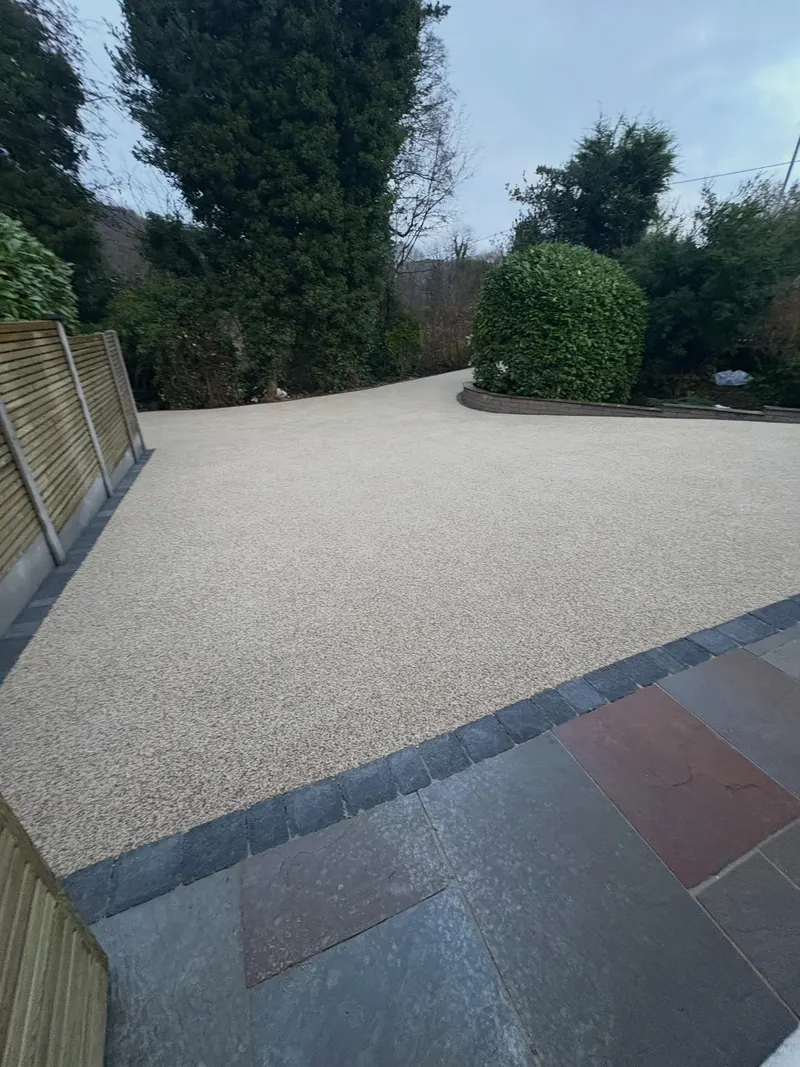 Expansive buff resin bound driveway curving away from a natural stone patio, separated by a dark block sett border and timber fencing.