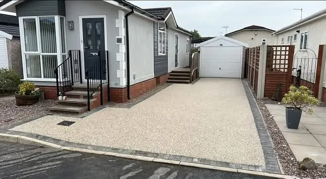 Light cream resin bound driveway with grey block borders between a grey-clad bungalow entrance and a white garage on an overcast day.