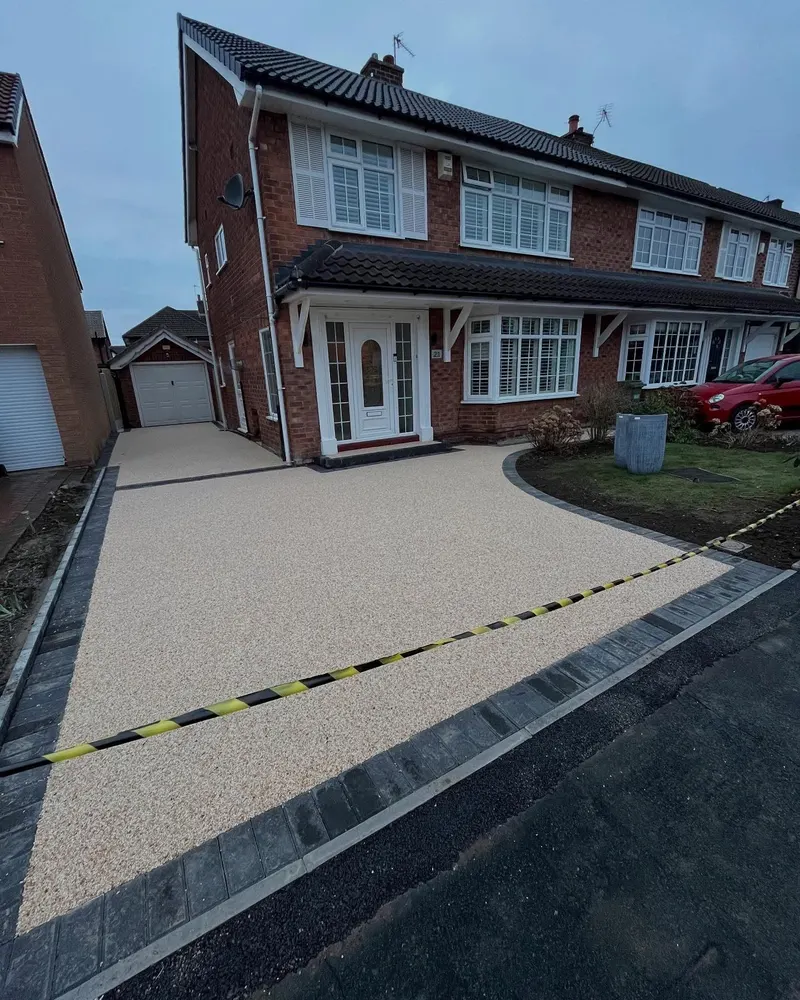 Buff resin bound driveway and side path with dark block borders in front of a red brick house, taped off while curing.