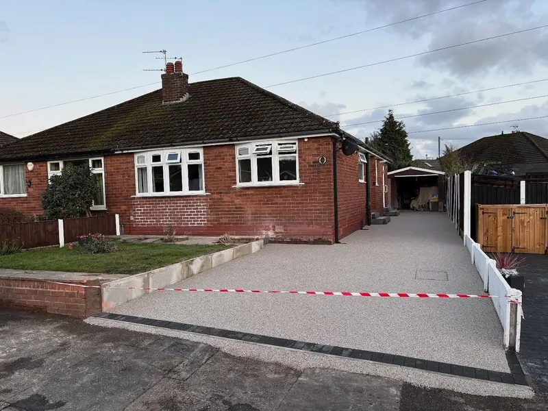 Light grey resin bound driveway with a front block border extending past a red brick bungalow toward a detached garage, with an integrated cover on the right.
