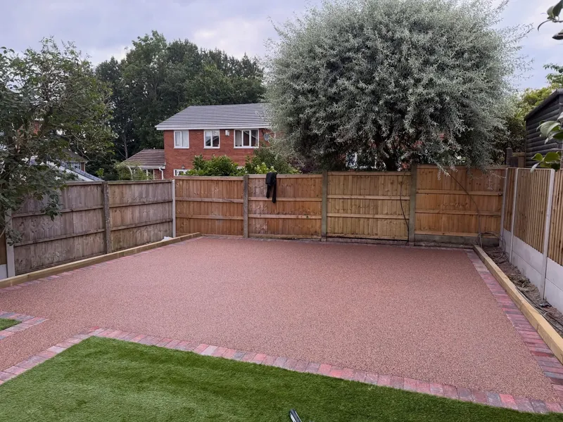 Earthy reddish resin bound patio rectangle bordered by mixed red and charcoal block pavers beside artificial grass and wooden fencing.