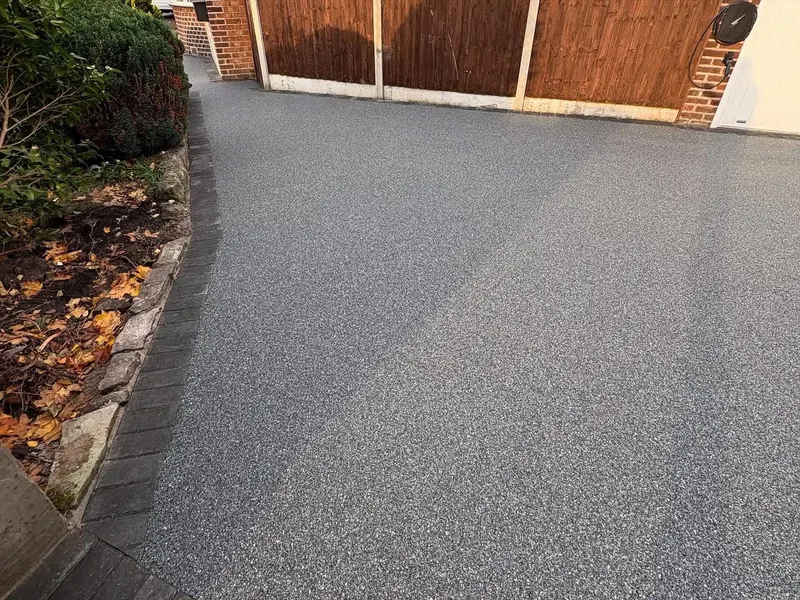 Mid-grey resin bound driveway with a curved charcoal block edge alongside shrubs, a wooden fence, and a brick wall with an EV charger.