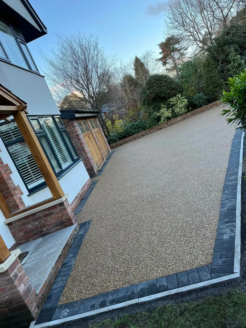 Large sandy-toned resin bound driveway framed by dark block paving in front of a white and brick house facing a small brick garage.